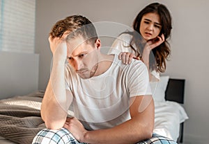 Young couple in the bedroom. Depressed man is sitting on the edge of bed while his girlfriend is trying to calm him down