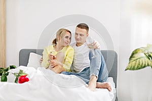A young couple in the bedroom with glasses of wine and flowers celebrating Valentine& x27;s Day or anniversary