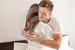 Young couple in the bedroom. Smiling unfaithful man is cheating and texting lover on the phone while hugging his