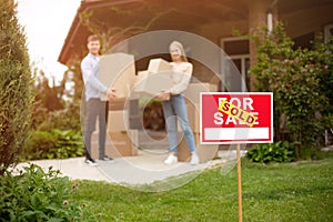 Young couple with boxes in front of their new house on moving day, focus on FOR SALE sign. Blank space