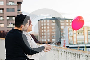 Young couple, boy and girl, with their backs turned and unrecognizable, holding a red heart-shaped balloon on a balcony with the
