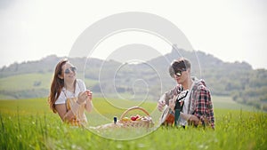A young couple in bright clothes sitting on a bright green meadow and having a picnic - a man playing guitar and woman