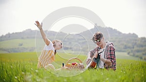 A young couple in bright clothes sitting on a bright green meadow - a man playing guitar while his happy girlfriend