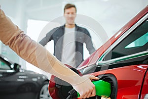 Young couple buying first electric car in the showroom. Close-up of hand charging electric car with the power cable