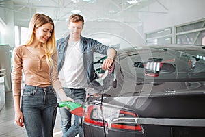 Young couple buying first electric car in the showroom. Woman charging ecological hybrid car with the power cable supply