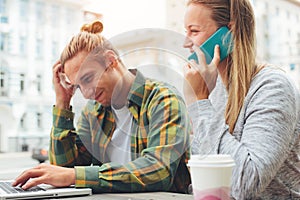 Young couple in cafe with laptop and mobile phone, confused man and woman talking on the phone