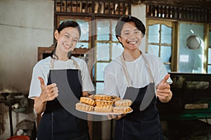 young couple of cake makers wearing aprons with thumbs up as a plate of baked cookies