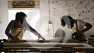 A young couple of carpenter working together as team in a carpentry workshop. A man cutting with a piece of timber with an