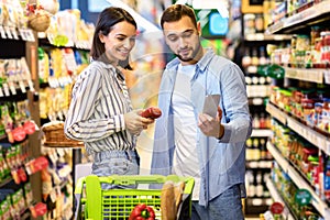 Young couple with cart and smartphone shopping in supermarket