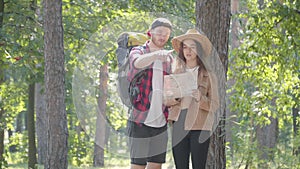 Young couple of Caucasian hikers walking in sunny forest, looking at paper map, and leaving. Portrait of confident