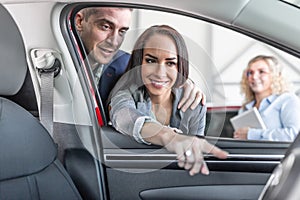 Young couple checks the features of new car interior while the sales manager stands aside in teh car dealership