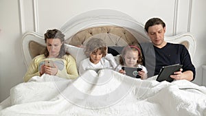 Young Couple with Children are Lying on the Bed Holding in Hands Electronic Gadgets and Talking to Each Other. Parents