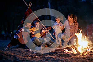 A young couple and children playing guitar around a campfire in the forest