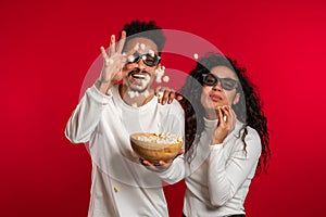 Young couple in cinema in 3d glasses watching fascinating comedy movie and eating popcorn on red studio background.
