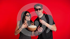 Young couple in cinema in 3d glasses watching fascinating horror movie and eating popcorn on red studio background.