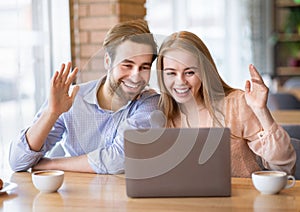 Young couple communicating online on laptop at cafe, waving to camera