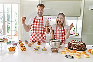 Young couple cooking pastries at the kitchen screaming proud, celebrating victory and success very excited with raised arms