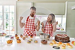 Young couple cooking pastries at the kitchen very happy and excited doing winner gesture with arms raised, smiling and screaming