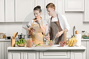 young couple cooking salad and reading recipe in cookbook