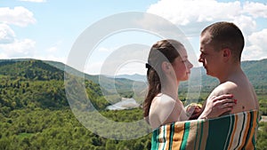 Young couple covered in lined blanket holding each other on the top of the hill with amazing forest hill view