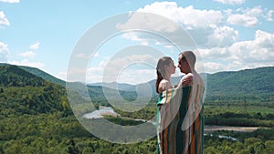 Young couple covered in lined blanket holding each other on the top of the hill with amazing view