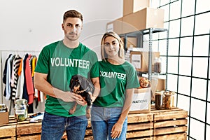 Young couple with cute dog wearing volunteer t shirt at donations stand thinking attitude and sober expression looking self