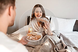 Young couple with delicious breakfast in the bed. Beautiful woman is eating fresh croissant with juice
