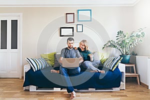 Young couple doing shoppings online on the sofa at home, looking at laptop screen and smiling