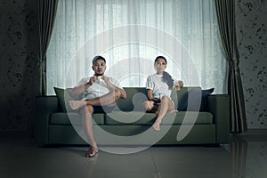 A young couple dressed casually sits on a green couch against a curtained background, gazing pensively