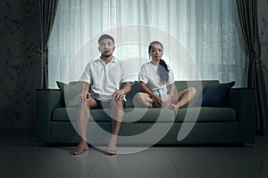 A young couple dressed casually sits on a green couch against a curtained background, gazing pensively