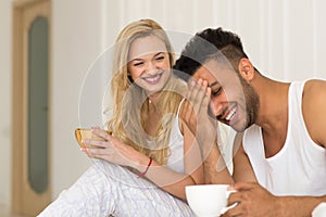 Young Couple Drink Coffee Sitting In Bed Laughing, Happy Smile Hispanic Man And Woman