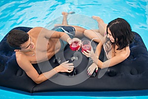 Young couple drinking cocktails on a mattress in the swimming pool enjoying each other and a summer holiday, top view