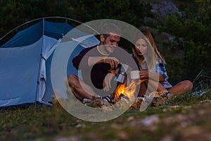 Young couple drinking coffee, enjoying the mountain view and warming up on the bonfire in the dusk