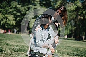 Young couple enjoying a playful moment in a sunny park, with the man sitting and woman leaning over him