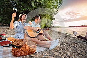 Young couple enjoying their time, having romantic picnic at the beach. Playing guitar and singing