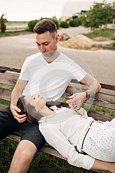 Young couple enjoying time together - woman lying on knees of man on bench on summer sunny day. Smiling man and woman looking on