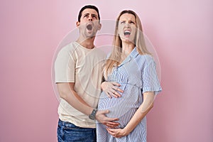 Young couple expecting a baby standing over pink background angry and mad screaming frustrated and furious, shouting with anger