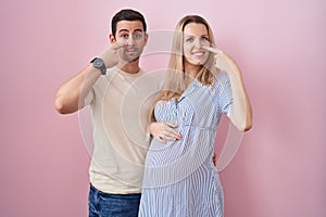 Young couple expecting a baby standing over pink background pointing with hand finger to face and nose, smiling cheerful