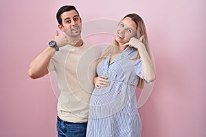 Young couple expecting a baby standing over pink background smiling doing phone gesture with hand and fingers like talking on the