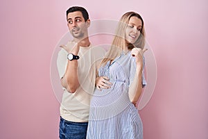 Young couple expecting a baby standing over pink background smiling with happy face looking and pointing to the side with thumb up