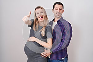 Young couple expecting a baby standing over white background approving doing positive gesture with hand, thumbs up smiling and