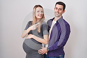 Young couple expecting a baby standing over white background cheerful with a smile on face pointing with hand and finger up to the