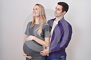 Young couple expecting a baby standing over white background looking away to side with smile on face, natural expression