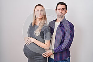 Young couple expecting a baby standing over white background puffing cheeks with funny face