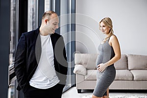 Young couple expecting baby standing together indoors near window. Young stylish family standing at home interior. Pragnancy