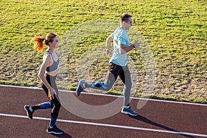 Young couple of fit sportsmen boy and girl running while doing exercise on red tracks of public stadium outdoors