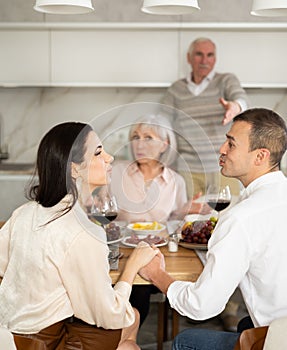 Young couple flirting sitting around table together with old family members looking with objection