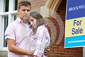 Young Couple Forced To Sell Home Through Financial Problems Standing Outdoors Next To For Sale Sign