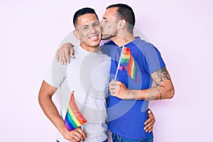 Young couple of gay men together in love holding homosexual pride colorful flag kissing with affection