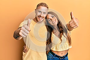 Young couple of girlfriend and boyfriend hugging and standing together approving doing positive gesture with hand, thumbs up